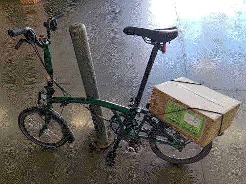 zelený Brompton C opřený o sloupek s naloženou kartonovou krabicí na zadním nosiči